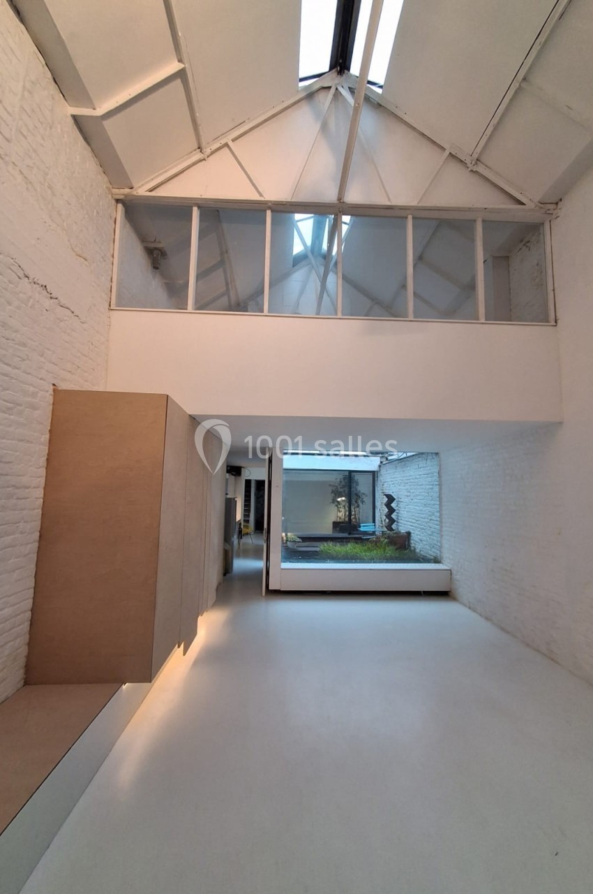 Intérieur lumineux avec murs blancs, verrière en hauteur et vue sur un patio avec végétation.