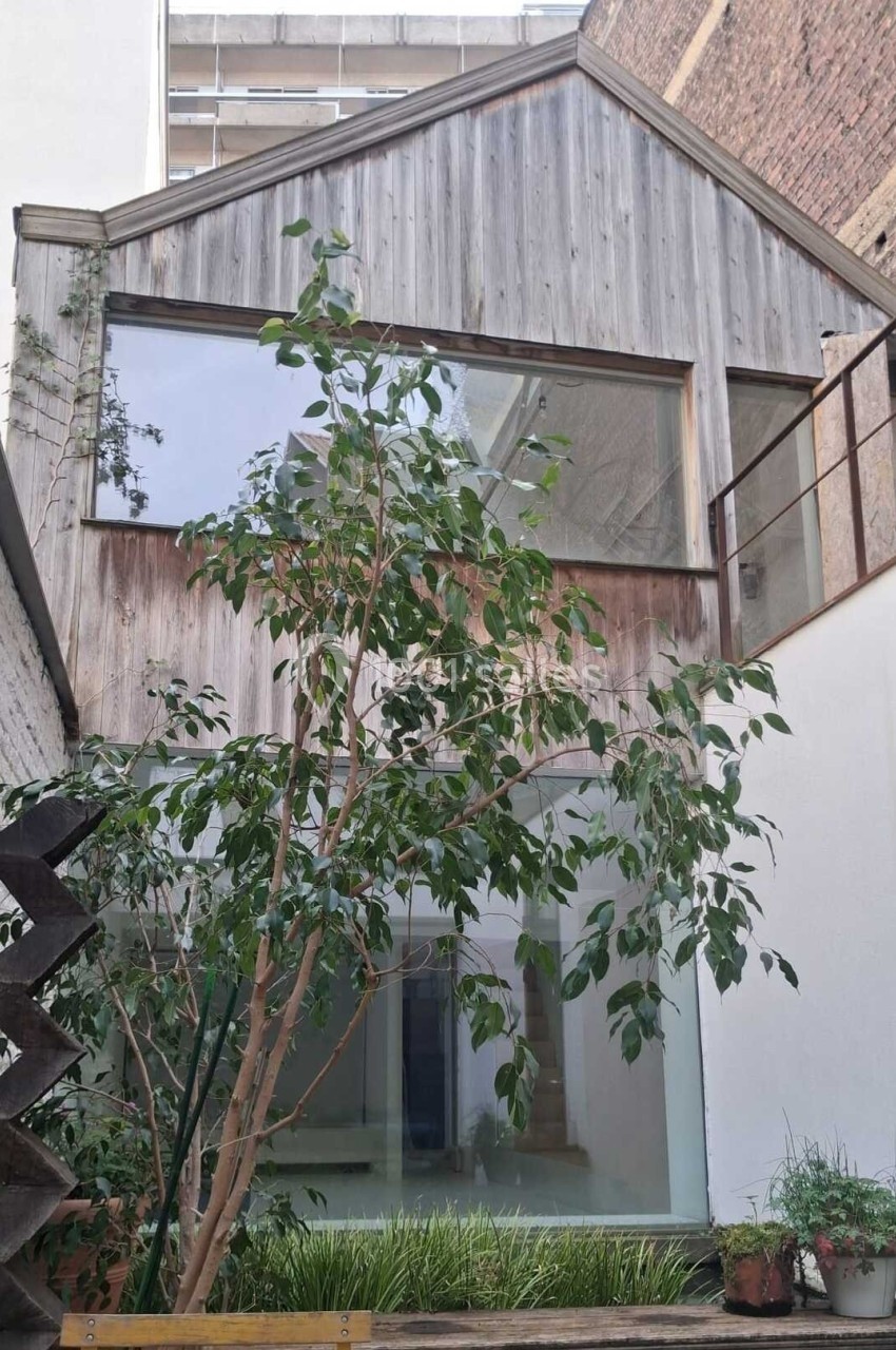 Façade d'une maison moderne en bois avec de grandes baies vitrées, entourée de murs en briques et végétation.
