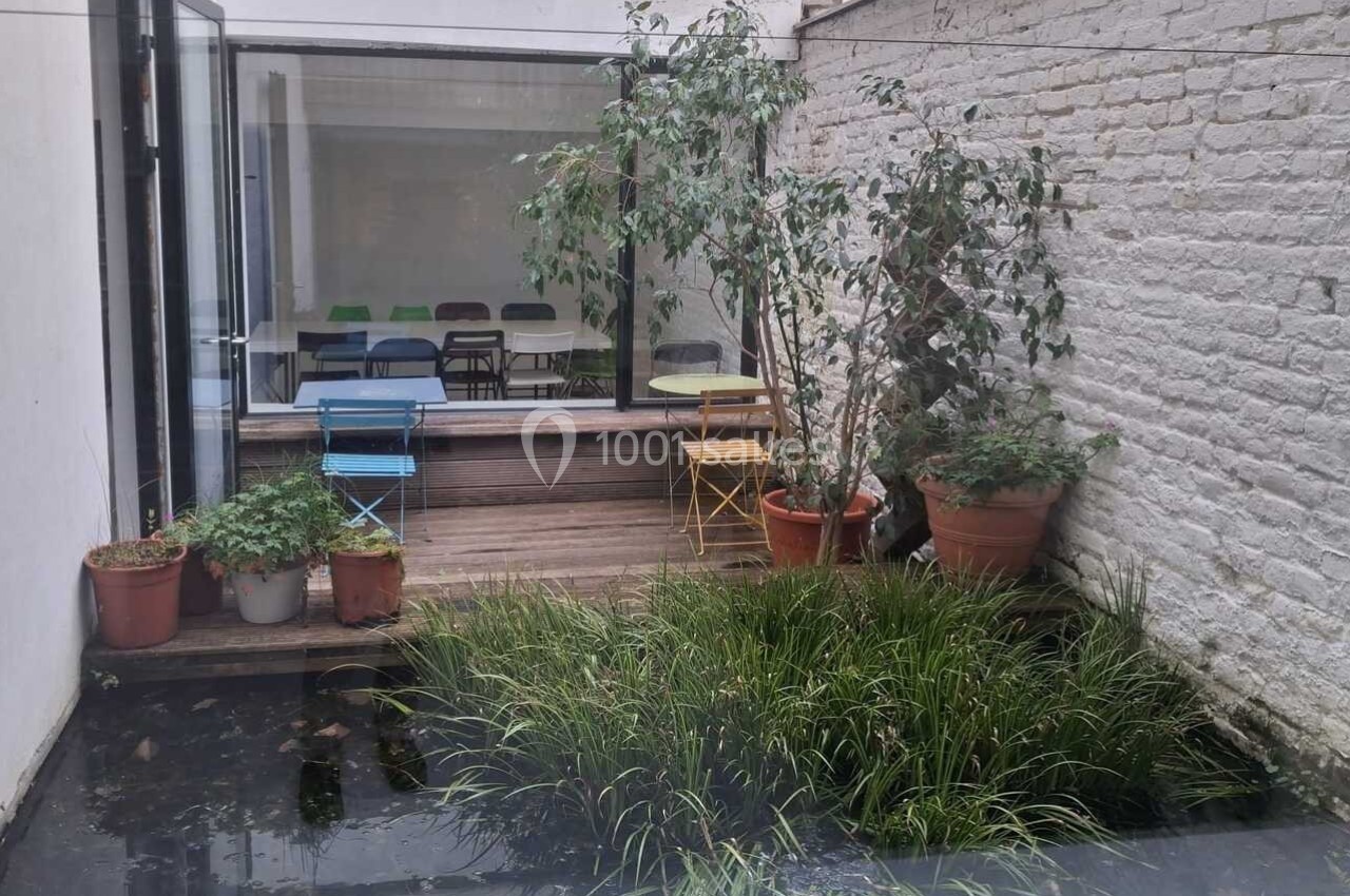 Petit jardin avec bassin, plantes en pots, et terrasse en bois, donnant sur une salle avec des chaises colorées.