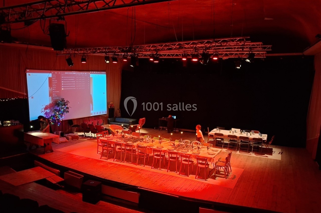 Salle de spectacle éclairée en rouge avec une grande table dressée, des chaises, un écran projeté et des équipements…