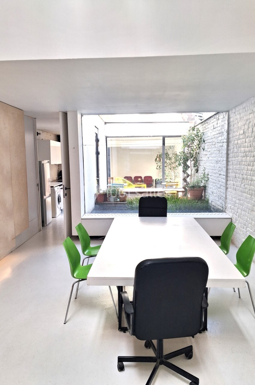 Salle de réunion lumineuse avec table blanche, chaises vertes et vue sur une cour intérieure avec plantes.