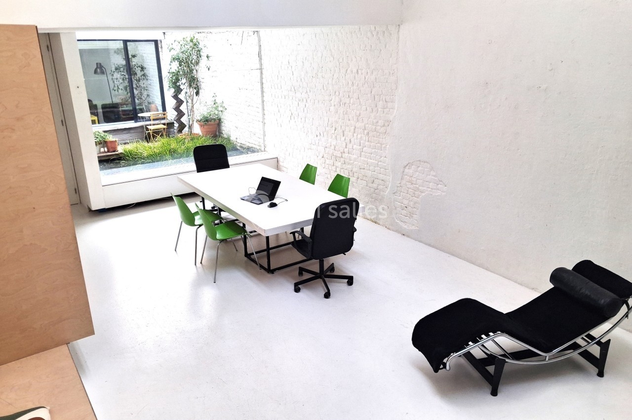 Salle de réunion lumineuse avec table blanche, chaises vertes et noires, et vue sur une cour intérieure avec plantes.
