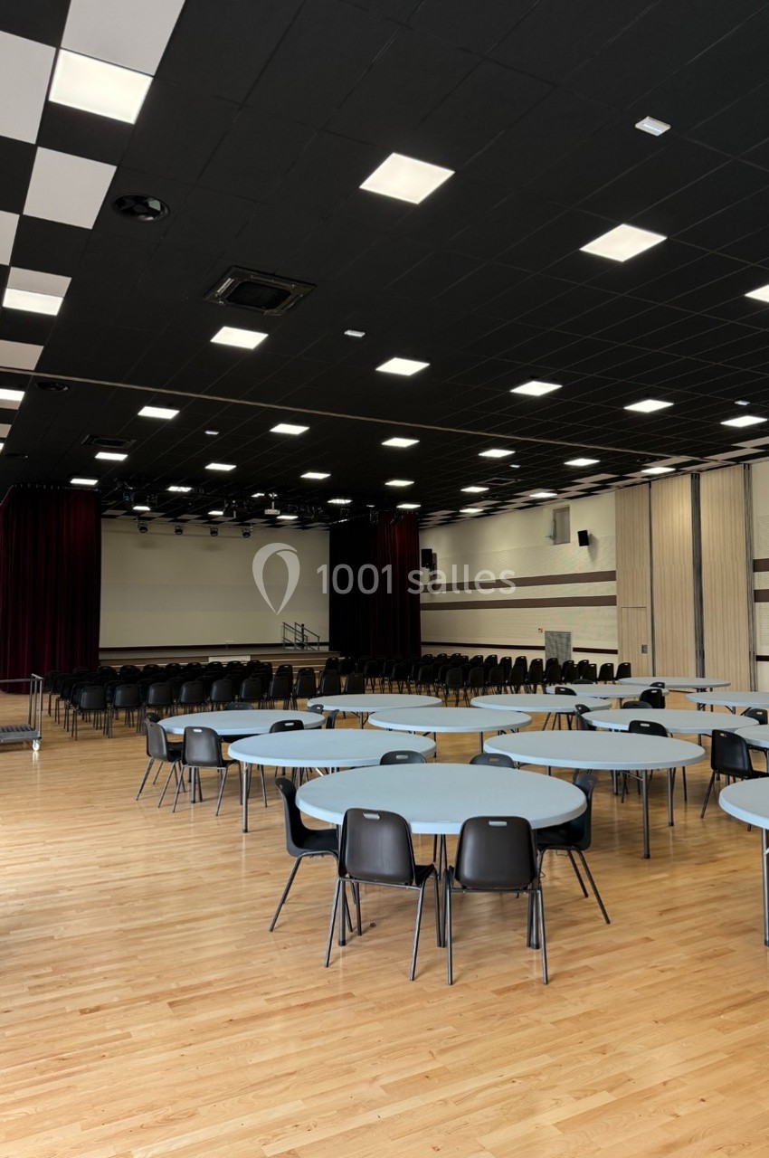 Salle polyvalente avec parquet, tables rondes, chaises noires et scène en fond, éclairée par des plafonniers.