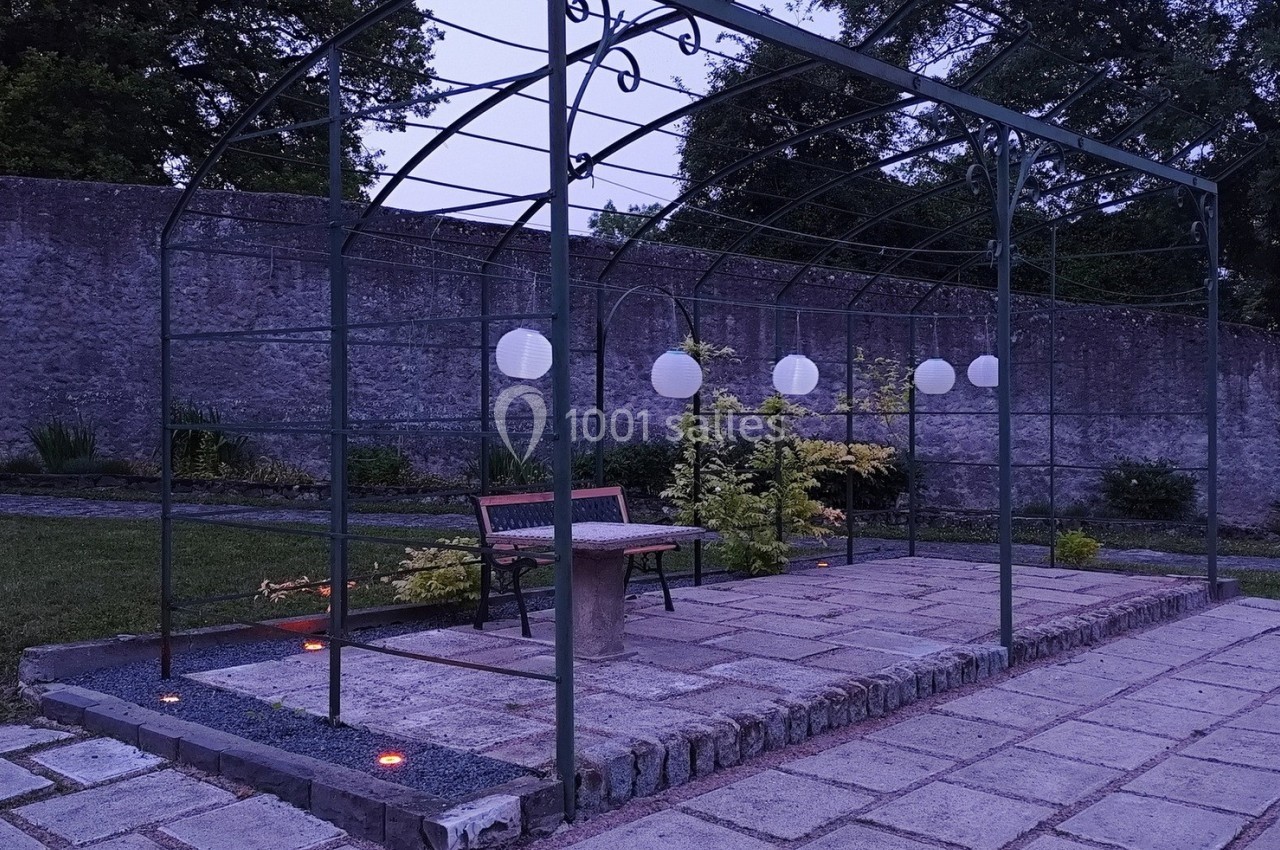 Pergola métallique dans un jardin, éclairée par des lampes suspendues et des lumières au sol au crépuscule.