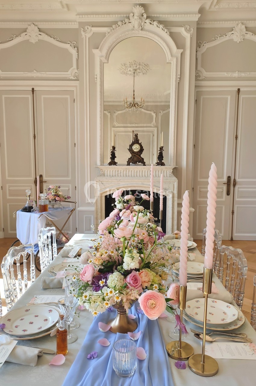 Table élégamment dressée avec fleurs pastel, chandeliers roses torsadés et vaisselle raffinée dans une salle classique.