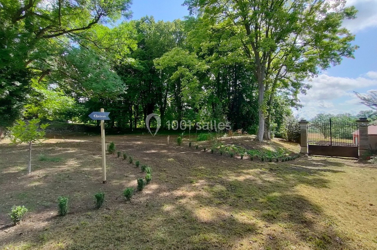Allée bordée de jeunes arbustes menant à une porte en fer, entourée d'arbres et d'un espace herbeux.