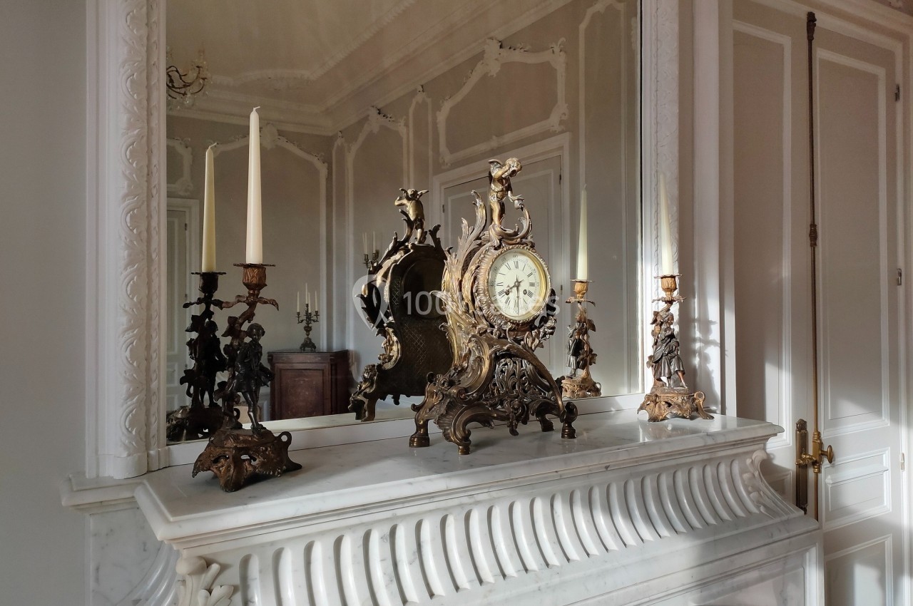 Horloge ancienne en bronze doré entourée de chandeliers sur une cheminée en marbre blanc, reflétée dans un miroir.