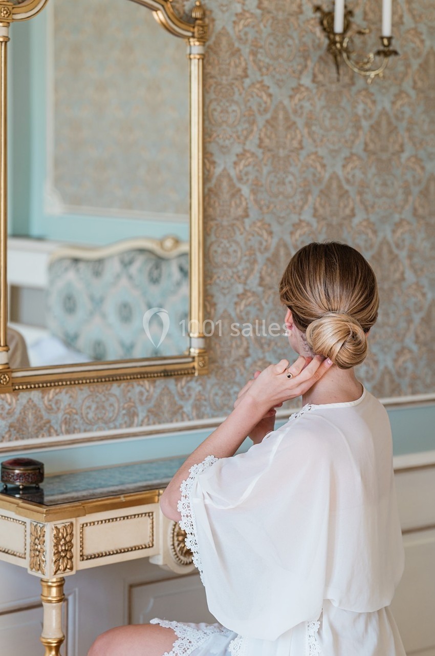 Femme assise devant une coiffeuse dorée, ajustant son chignon, dans une pièce élégante au décor classique.