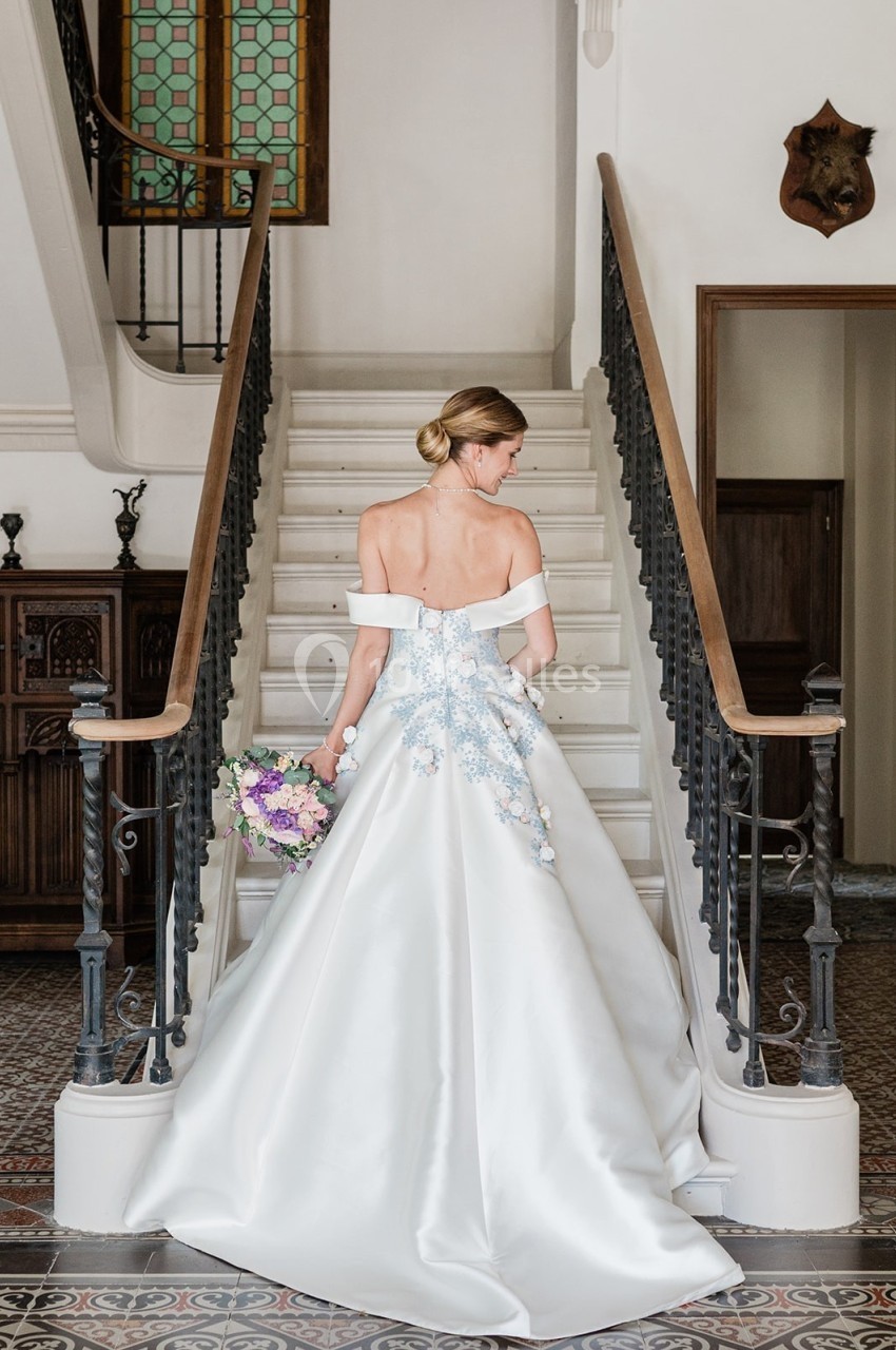 Femme en robe de mariée blanche avec détails floraux, tenant un bouquet, debout devant un escalier intérieur.