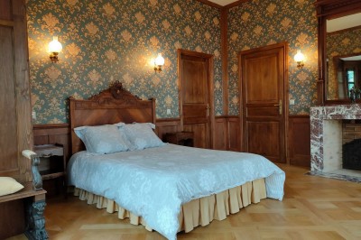 Miniature Chambre Bleue Allée bordée de jeunes arbustes menant à une porte en fer, entourée d'arbres et d'un espace herbeux.