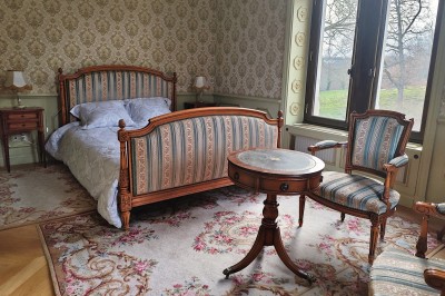 Miniature Chambre Dorée Allée bordée de jeunes arbustes menant à une porte en fer, entourée d'arbres et d'un espace herbeux.