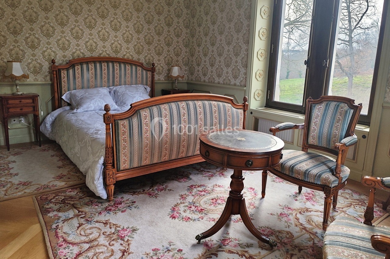 Chambre avec lit ancien, fauteuils assortis, table ronde en bois et grande fenêtre donnant sur un paysage verdoyant.