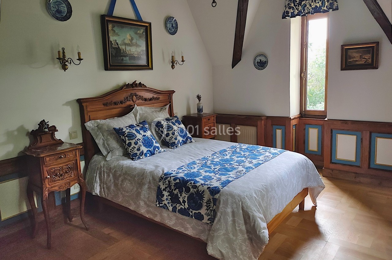 Chambre avec un lit double, linge de lit bleu et blanc, meubles en bois sculpté et une fenêtre laissant entrer la lumière.