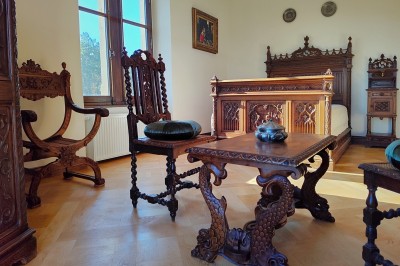 Miniature Chambre Gothique Allée bordée de jeunes arbustes menant à une porte en fer, entourée d'arbres et d'un espace herbeux.