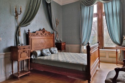 Miniature Chambre Turquoise Allée bordée de jeunes arbustes menant à une porte en fer, entourée d'arbres et d'un espace herbeux.