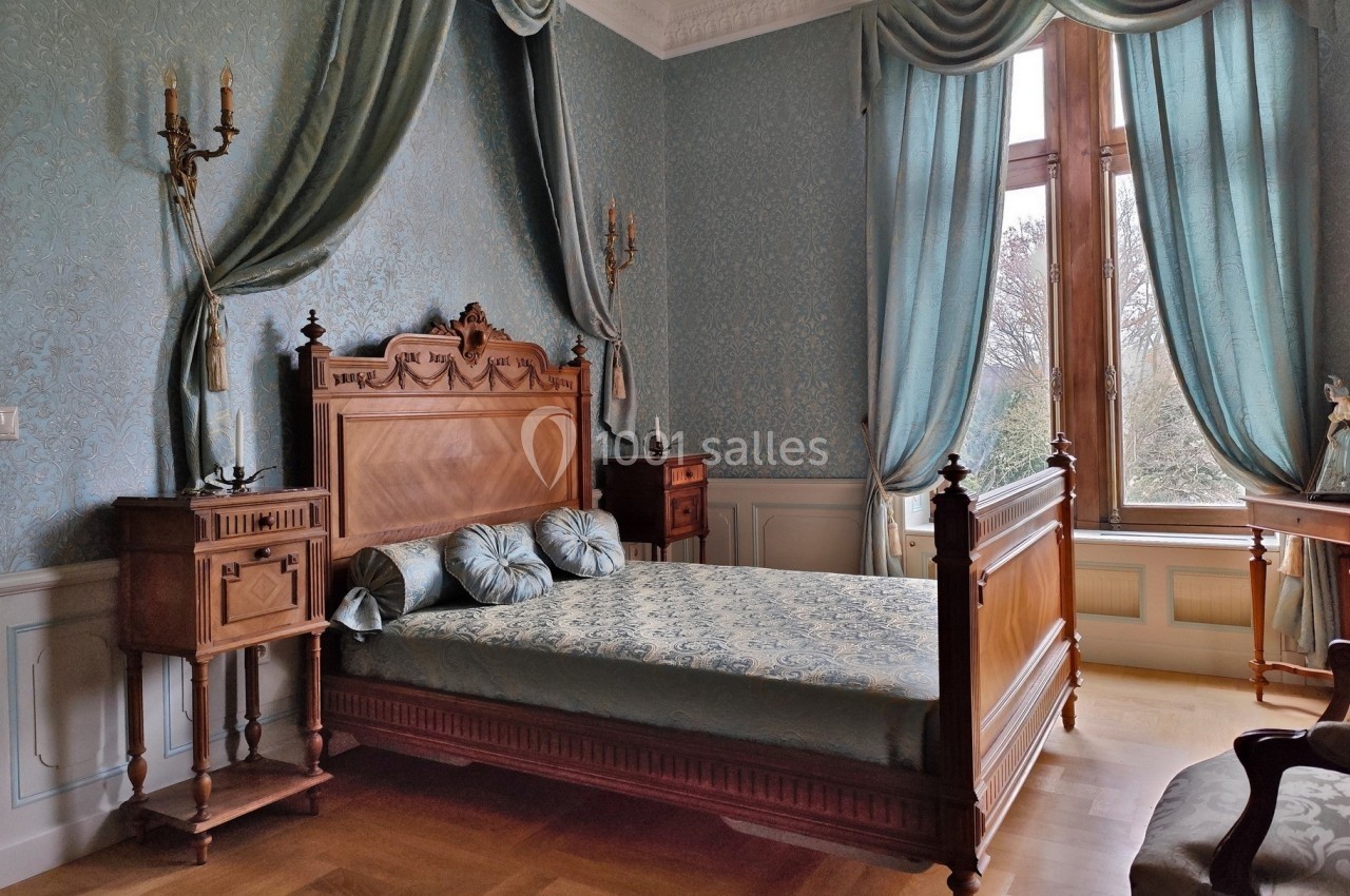 Chambre élégante avec lit en bois sculpté, rideaux et murs décorés de motifs classiques, éclairée par une grande fenêtre.