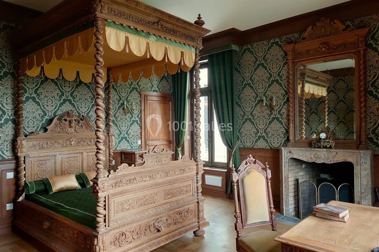 Chambre ancienne avec lit à baldaquin sculpté, cheminée, miroir et décorations murales à motifs verts.