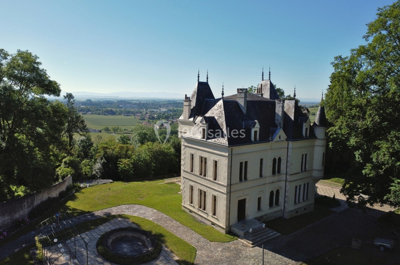 Vue aérienne d'un château entouré de verdure avec un paysage vallonné en arrière-plan sous un ciel dégagé.