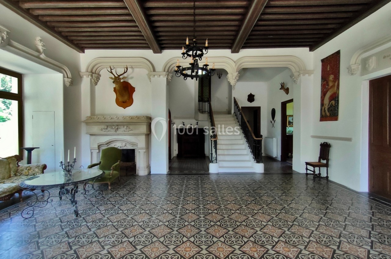 Salle avec sol en carreaux décoratifs, cheminée ornée, escalier en bois, et plafond à poutres apparentes.