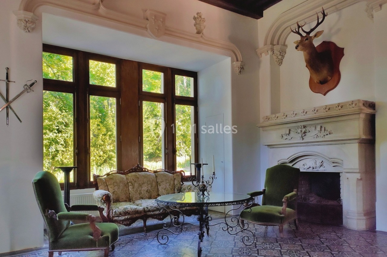 Salon élégant avec cheminée sculptée, tête de cerf, mobilier ancien et grande baie vitrée donnant sur un jardin.