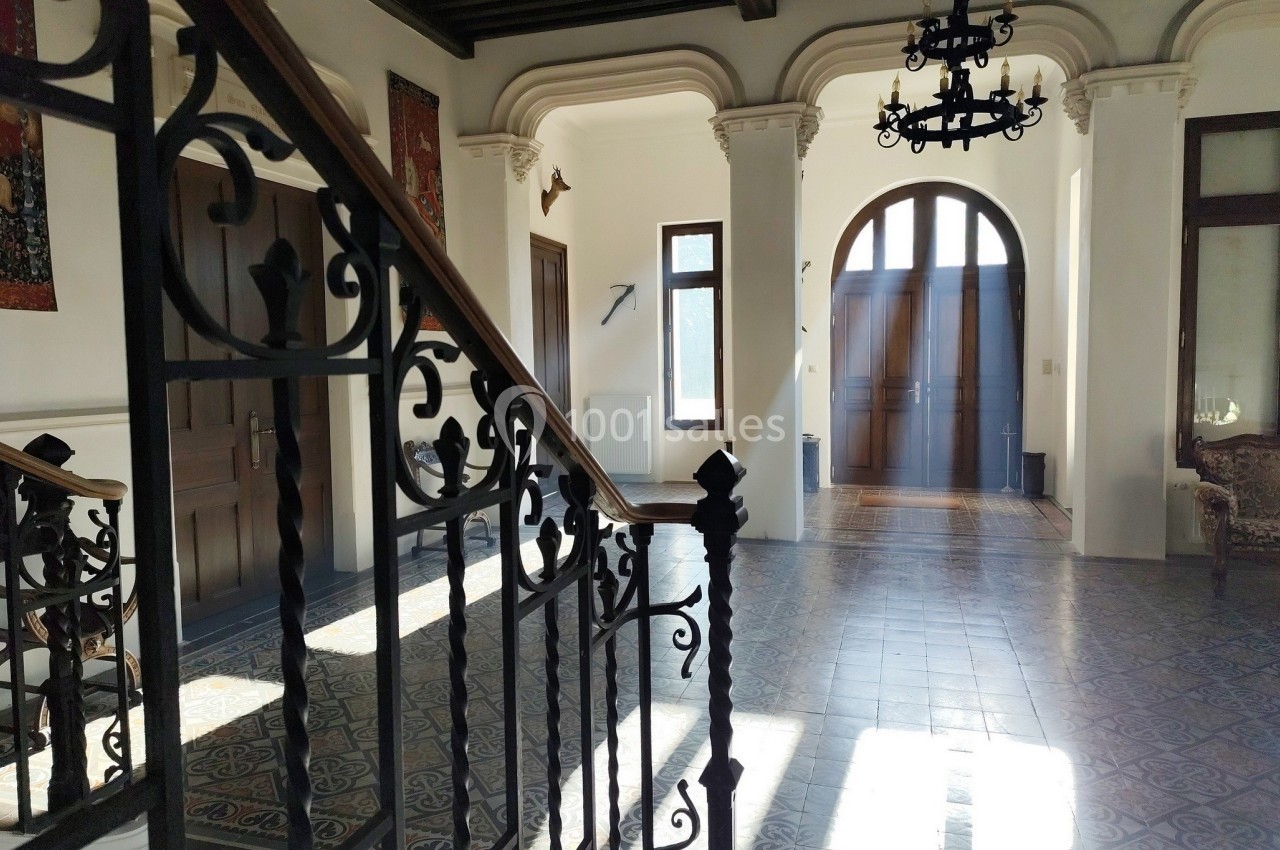 Escalier en fer forgé dans un hall lumineux avec carrelage ancien, portes en bois et lustres suspendus.