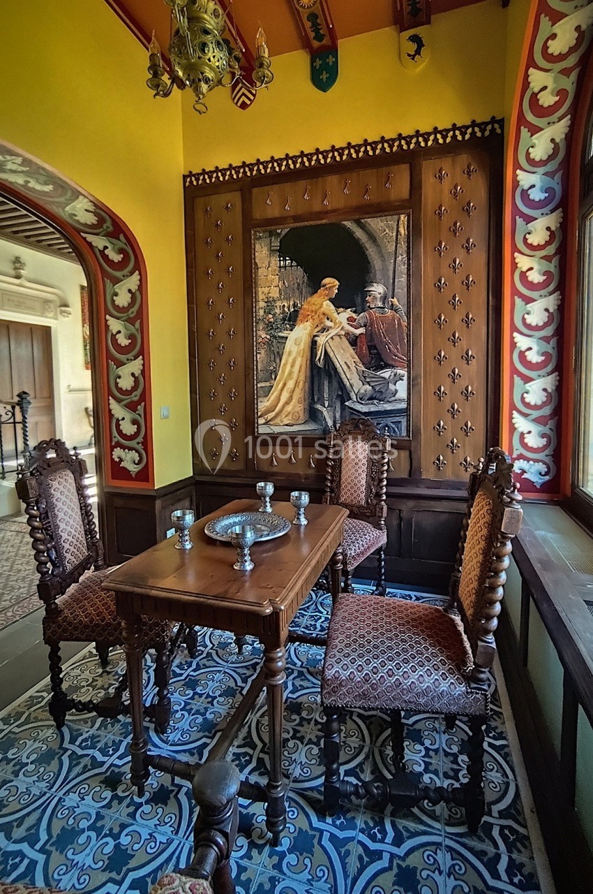 Salle décorée de style médiéval avec une table en bois, quatre chaises sculptées et un tableau représentant une scène…