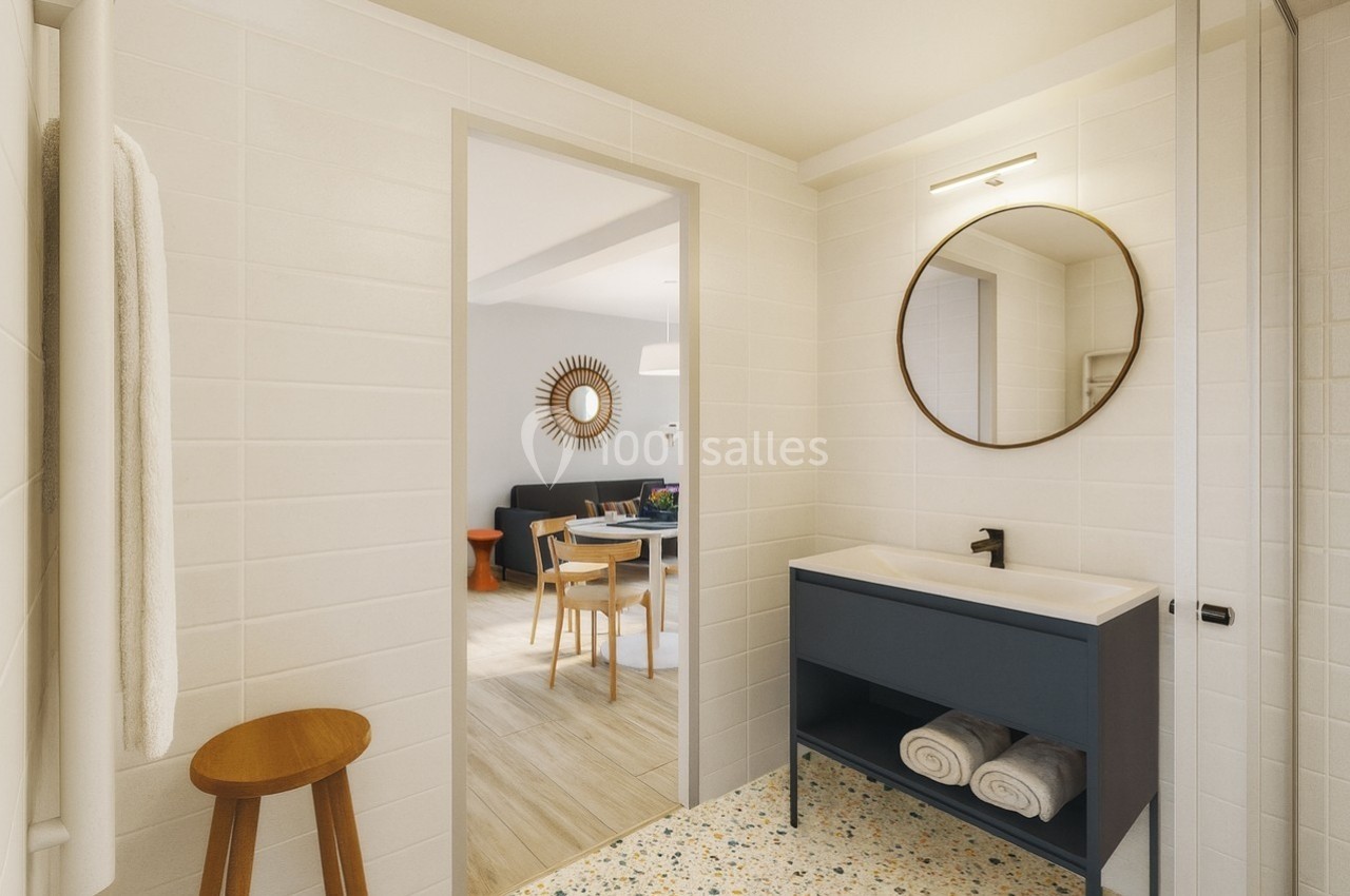 Salle de bain moderne avec meuble vasque bleu, miroir rond, et vue sur une salle à manger lumineuse en arrière-plan.