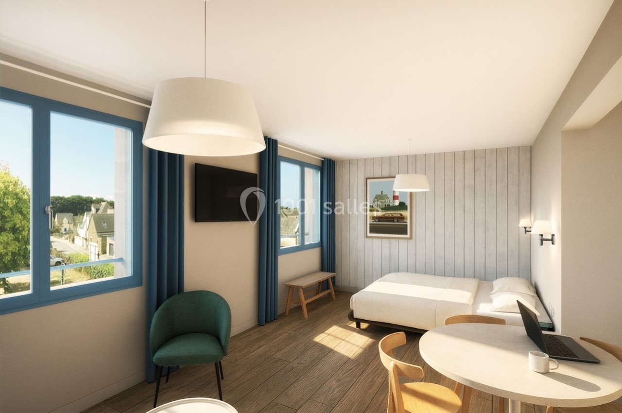 Chambre lumineuse avec lit double, table ronde, fauteuil, télévision murale et grandes fenêtres donnant sur l'extérieur.