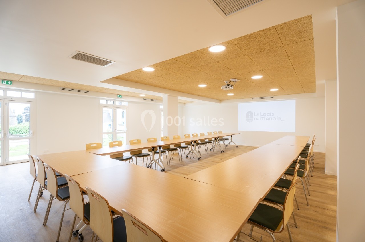 Salle de réunion lumineuse avec tables disposées en U, chaises en bois clair et projecteur au plafond.