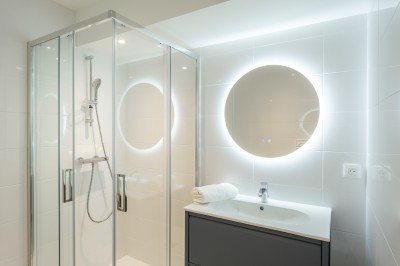 Salle de bain moderne avec douche vitrée, lavabo sur meuble noir et miroir rond éclairé.