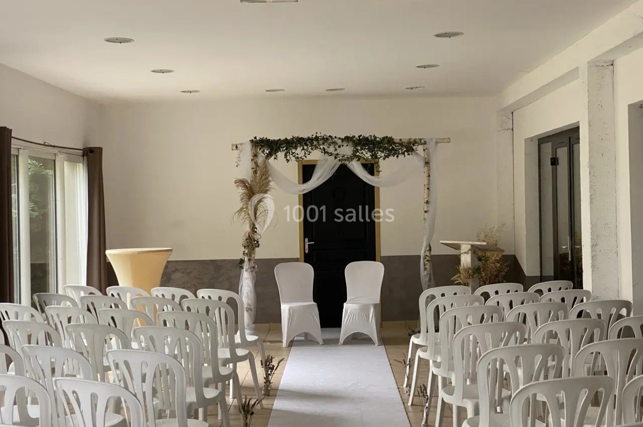 Salle de cérémonie décorée avec des chaises blanches alignées, une arche ornée de feuillage et un tapis blanc central.