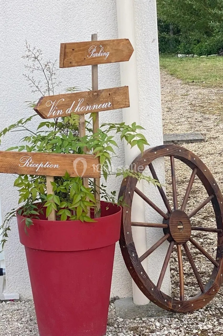 Panneaux en bois indiquant parking, vin d'honneur et réception, posés dans un pot rouge près d'une roue en bois.