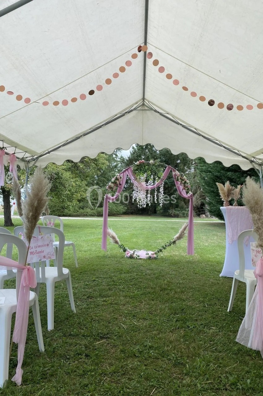 Tente blanche décorée pour une cérémonie en plein air, avec arche florale, chaises et rubans roses sur pelouse verte.