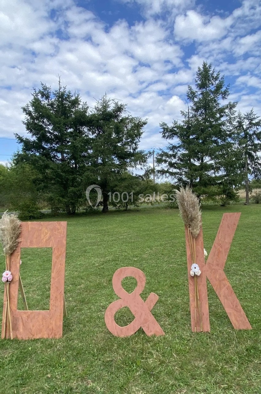 Lettres en bois ’D & K’ décorées de fleurs et de pampas, disposées sur une pelouse avec des arbres en arrière-plan.