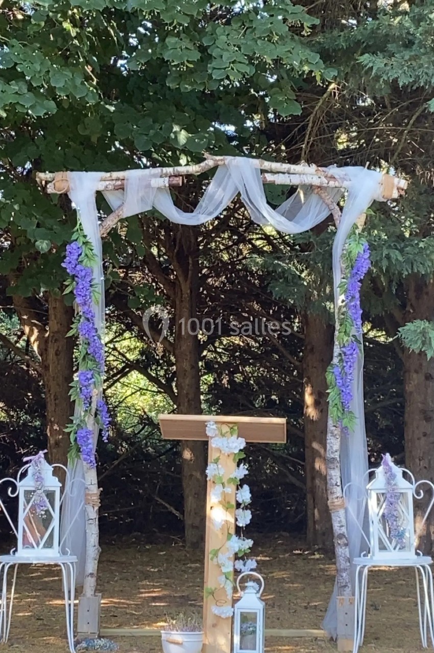 Arche de cérémonie décorée de voilages blancs, fleurs violettes et blanches, entourée de chaises blanches dans un jardin…