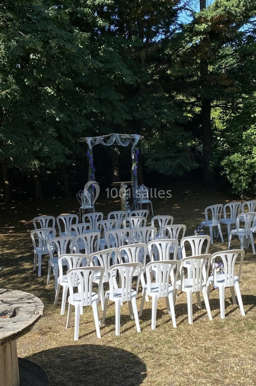 Chaises blanches disposées en rangées devant une arche décorée, dans un jardin ombragé.