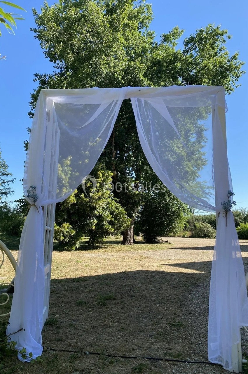 Arche décorée de voilages blancs installée en extérieur, entourée d'arbres sous un ciel dégagé.