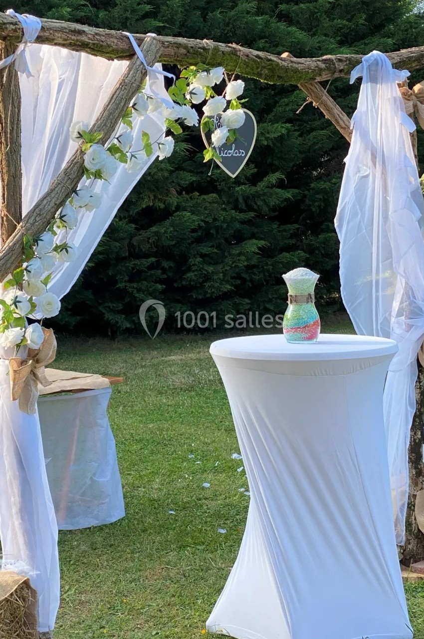 Arche en bois décorée de voilages blancs et de fleurs, avec une table haute drapée, dans un jardin verdoyant.
