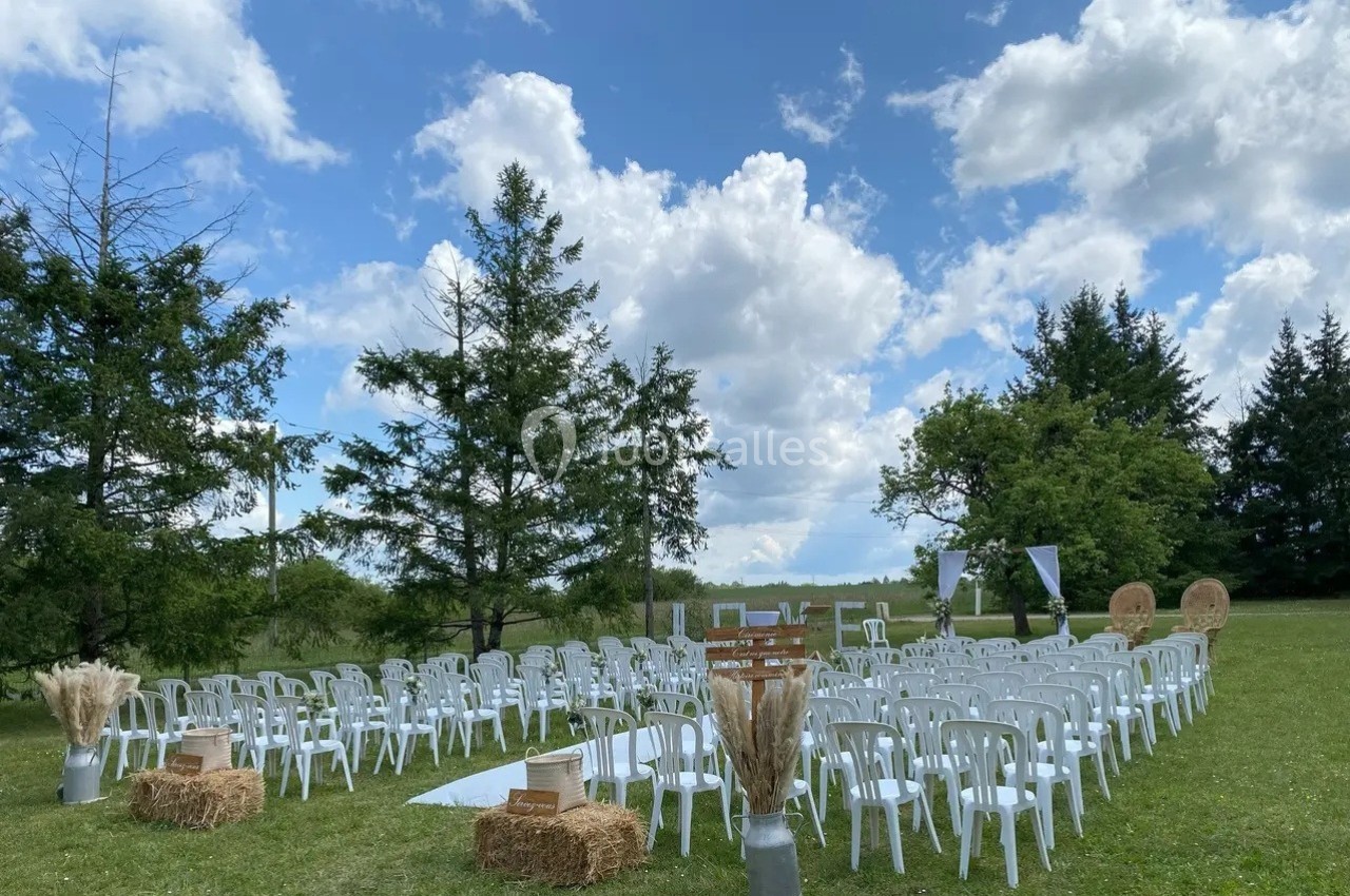 Chaises blanches disposées en extérieur pour une cérémonie, entourées d'arbres et sous un ciel partiellement nuageux.