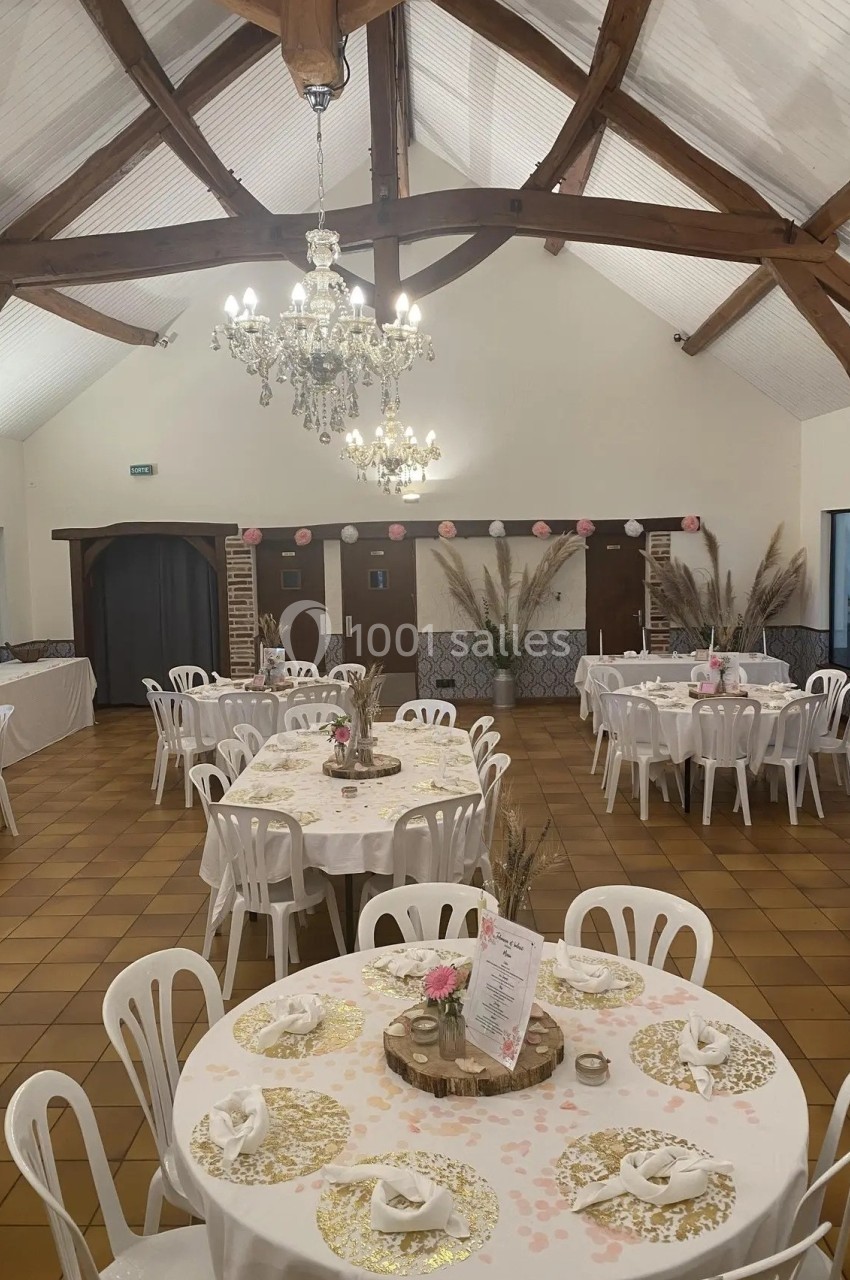 Salle de réception décorée avec des tables rondes, nappes blanches, centres de table floraux et chaises en plastique.