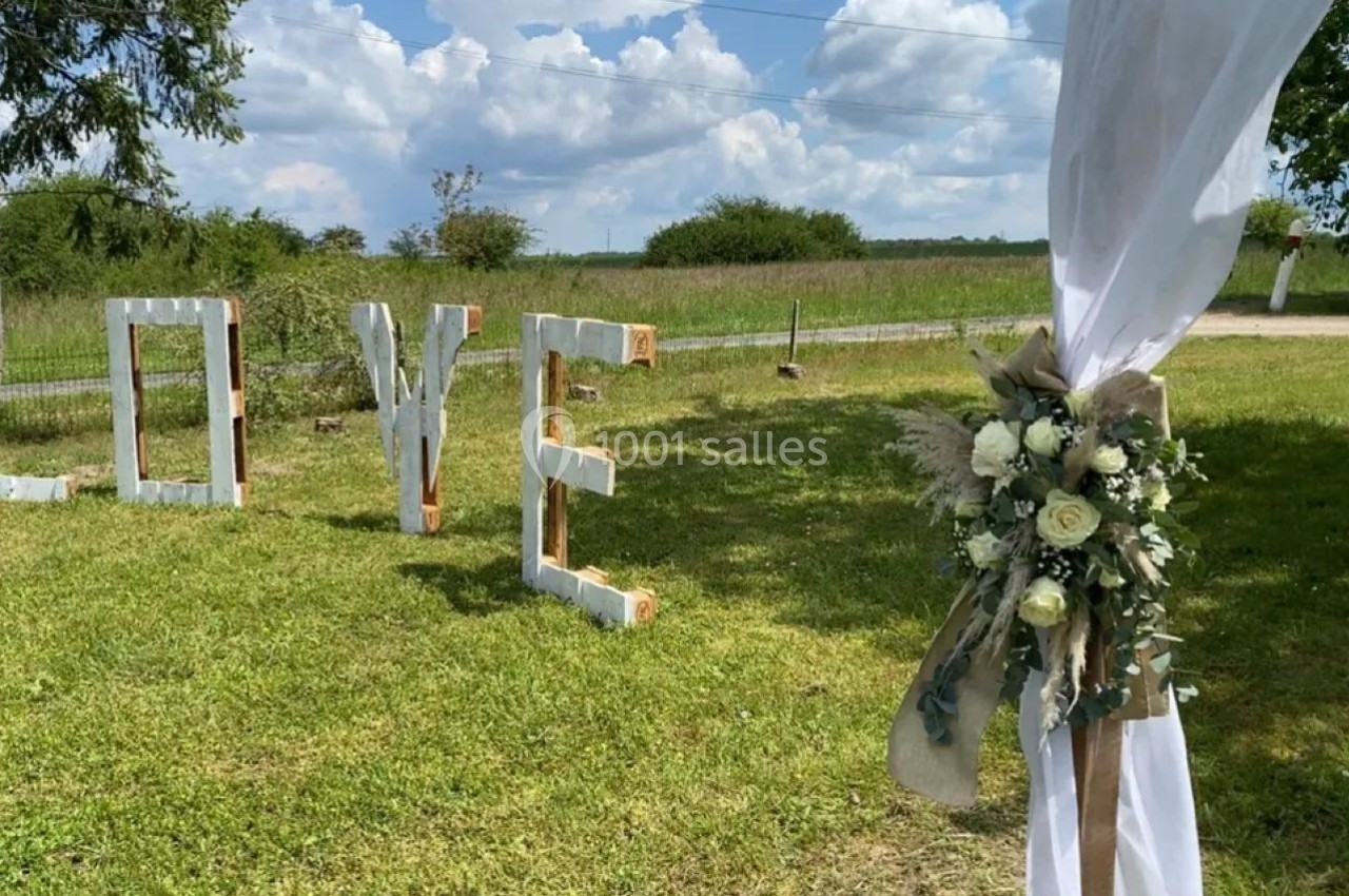 Lettres géantes formant ’LOVE’ sur une pelouse, avec un poteau décoré de fleurs blanches au premier plan.