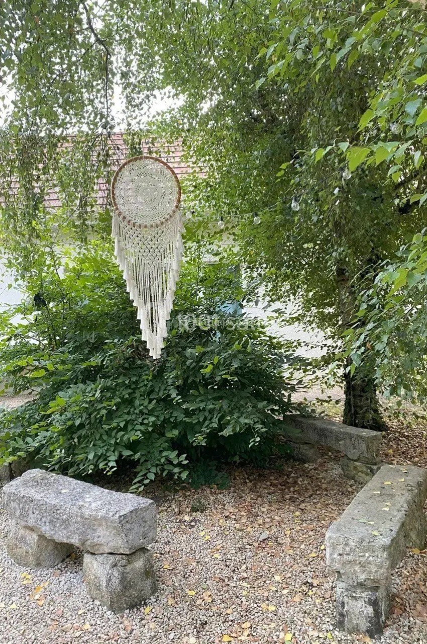 Attrape-rêves suspendu à une branche, entouré de verdure, avec des bancs en pierre et un sol couvert de feuilles.