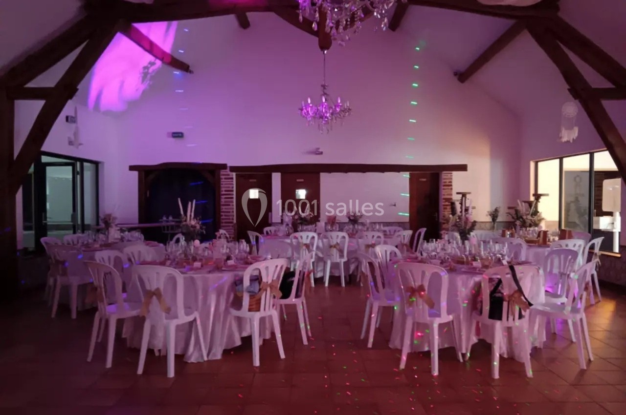 Salle de réception décorée avec des tables rondes, chaises blanches et éclairage coloré sous un plafond avec poutres…