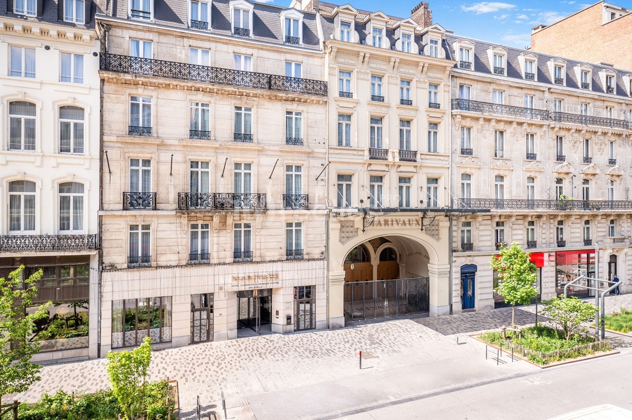 Façade d'immeubles haussmanniens avec une entrée voûtée et des commerces au rez-de-chaussée, rue pavée bordée d'arbres.