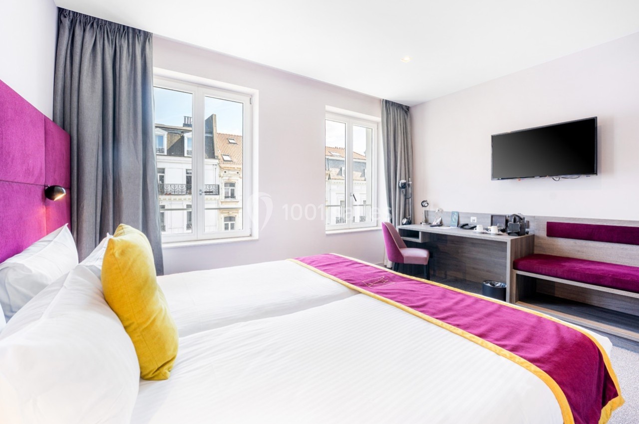Chambre lumineuse avec deux lits, draps blancs, coussins colorés, bureau, télévision murale et grandes fenêtres donnant sur…