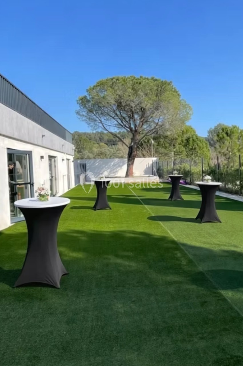 Tables hautes noires disposées sur une pelouse artificielle près d'un bâtiment moderne sous un ciel dégagé.