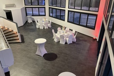 Salle avec tables rondes élégamment dressées, nappes blanches, chaises houssées et grandes fenêtres en arrière-plan.