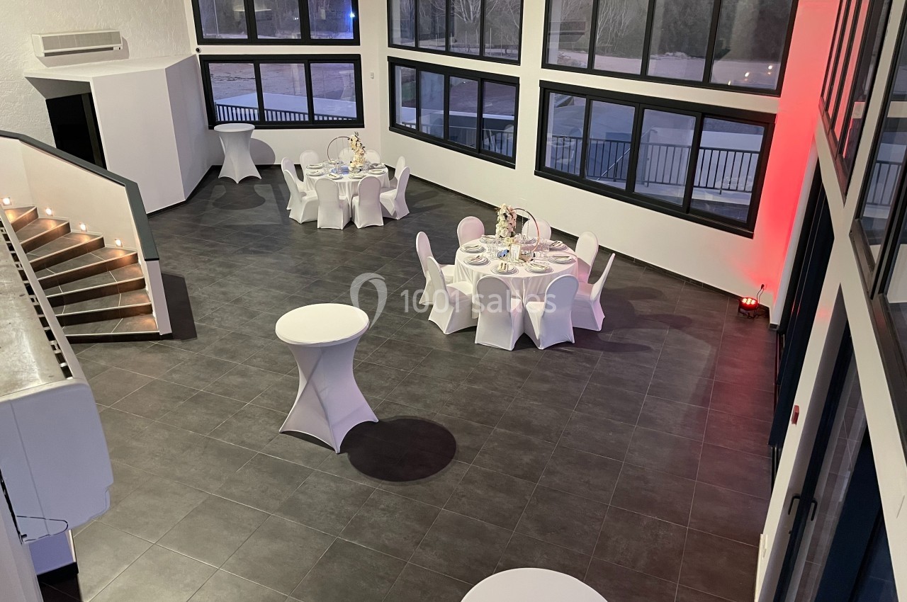 Salle de réception moderne avec tables rondes dressées, grandes baies vitrées et escalier éclairé.