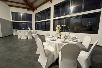 Salle avec tables rondes élégamment dressées, nappes blanches, chaises houssées et grandes fenêtres en arrière-plan.