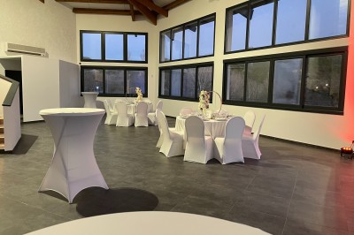 Salle avec tables rondes élégamment dressées, nappes blanches, chaises houssées et grandes fenêtres en arrière-plan.